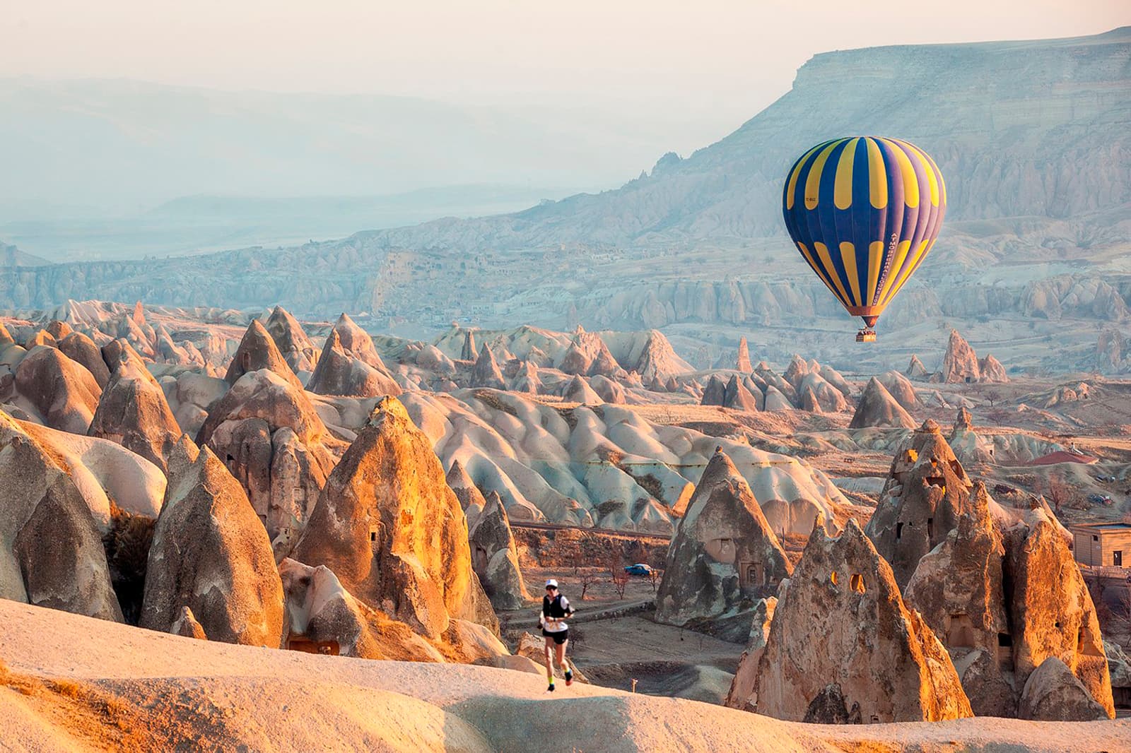 A person stands among unique rock formations in Cappadocia, Turkey, while a colorful hot air balloon floats above the landscape in soft, warm sunlight.