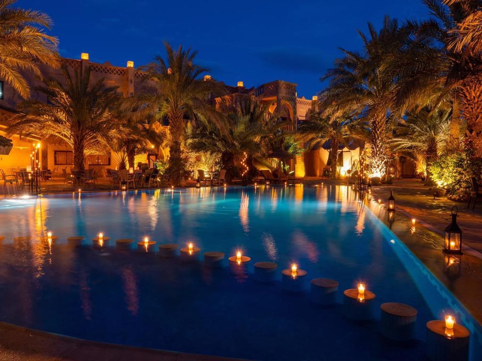 A serene pool illuminated by numerous scattered candles under a twilight sky, surrounded by tall palm trees and cozy, warm-lit buildings, creating a tranquil and inviting ambiance.