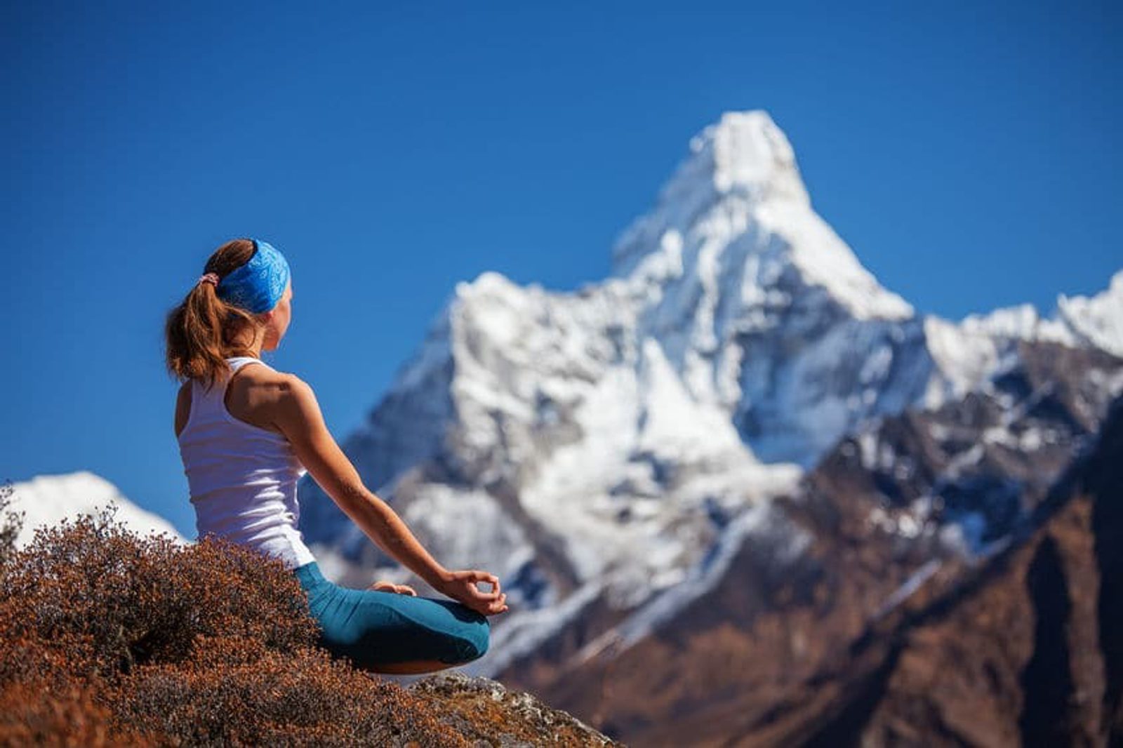 A person wearing a white tank top and blue pants sits in a meditative pose on a rocky surface. They face a snow-covered mountain under a clear blue sky, with a vivid backdrop of rugged terrains. The person wears a blue headband and their hair is tied back in a ponytail.