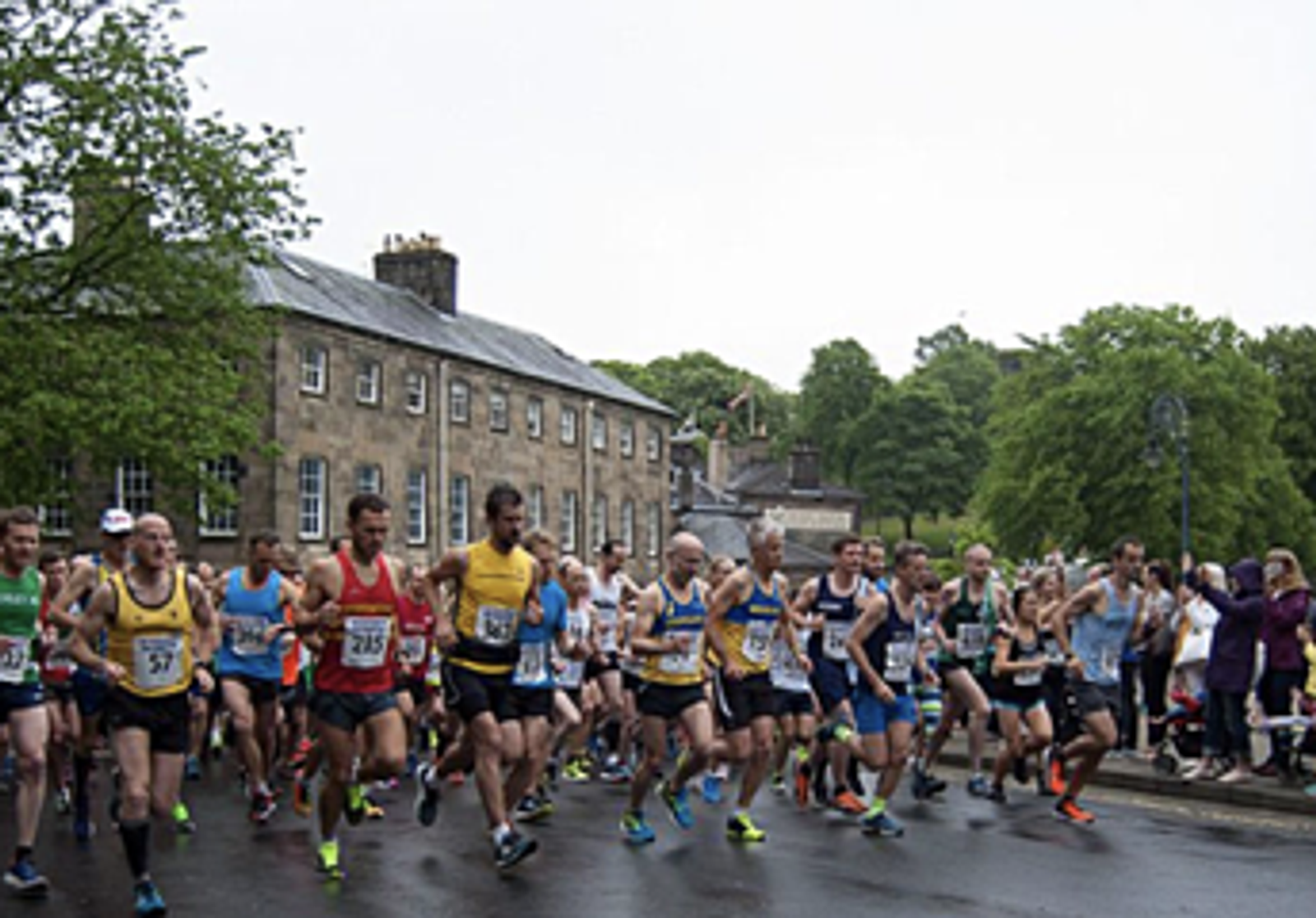 A large group of runners begins a race on a wet street, with many wearing colorful athletic clothing and numbered bibs. In the background, there are old stone buildings and lush green trees. The sky appears overcast, suggesting recent or impending rain.
