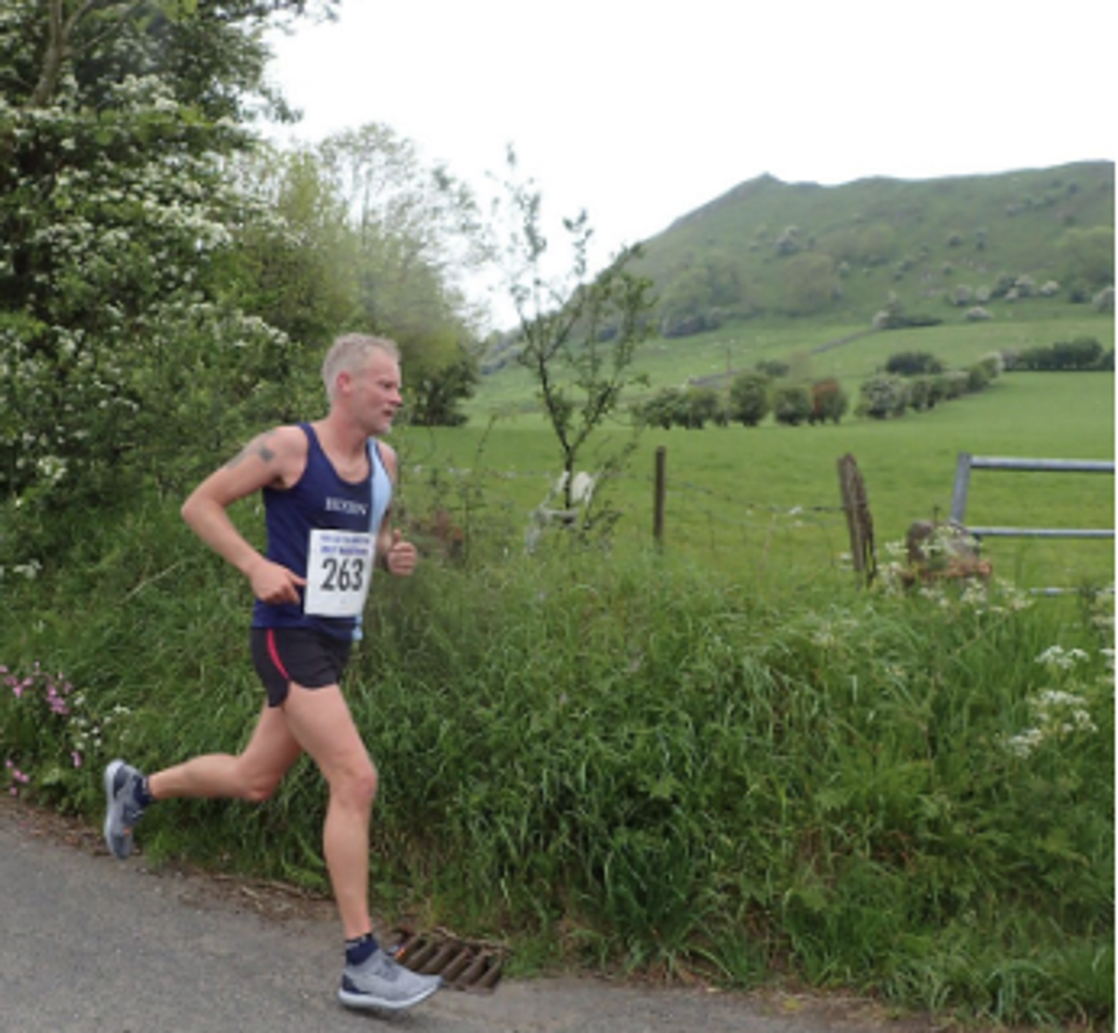 A man wearing a navy tank top with the number 263 and black shorts is running on a rural road. Green fields and hills with scattered trees are in the background, along with a fence and some bushes along the roadside.