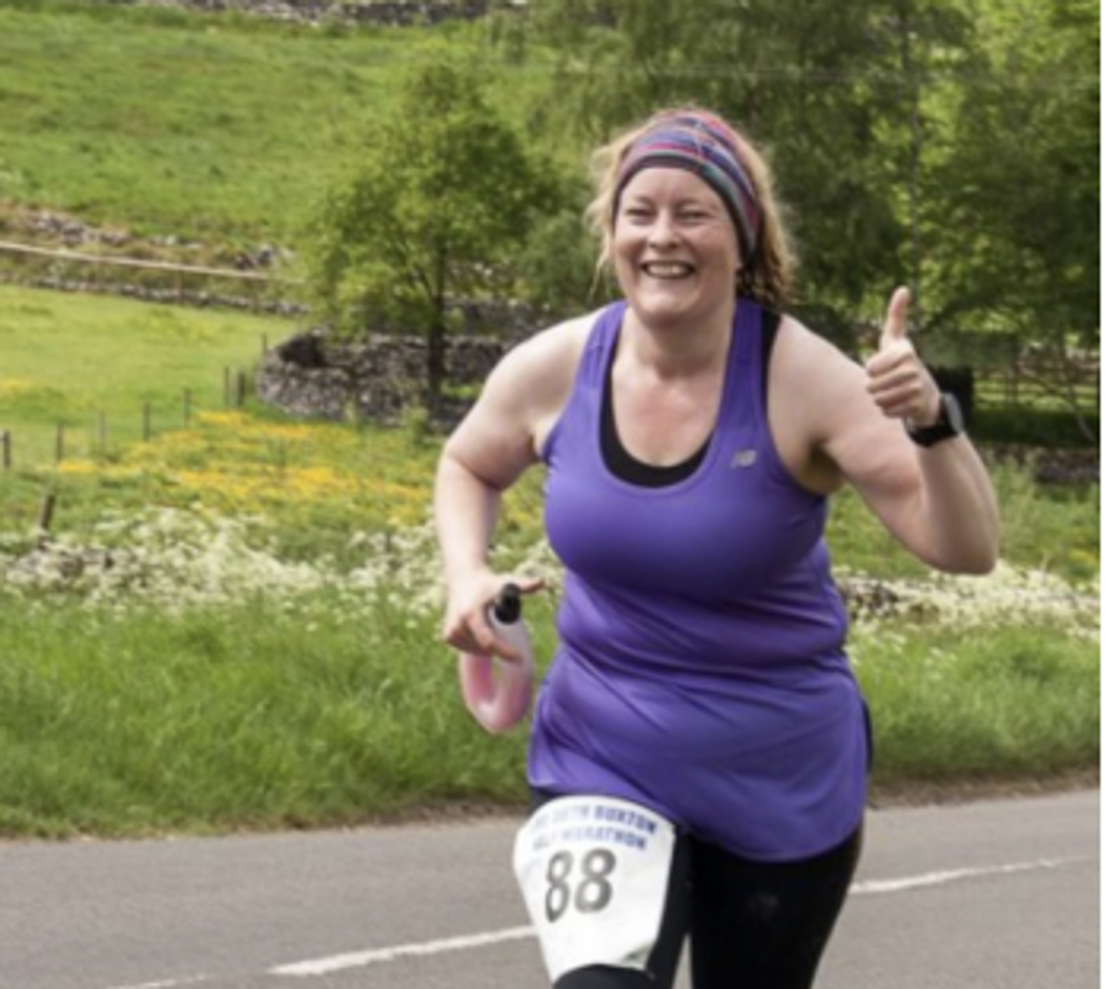 A woman in a purple tank top and black leggings is running outdoors, smiling broadly and giving a thumbs-up. She is wearing a race number "88" and a headband. The background shows a scenic countryside with greenery and a stone fence.