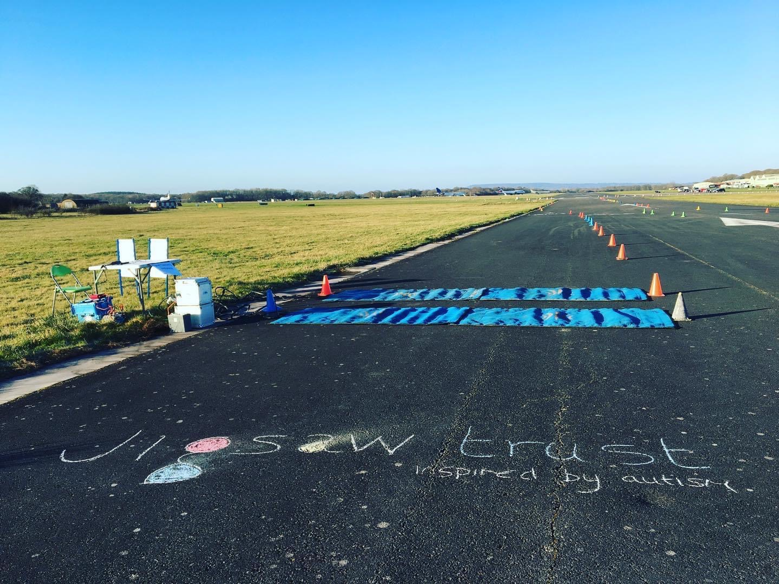 A runway decorated with colorful chalk drawings, banners, and signs that read "Jigsaw Trust inspired by autism." The path is lined with traffic cones, and there are tables and chairs set up alongside the runway on a grassy field. The sky is clear and blue.