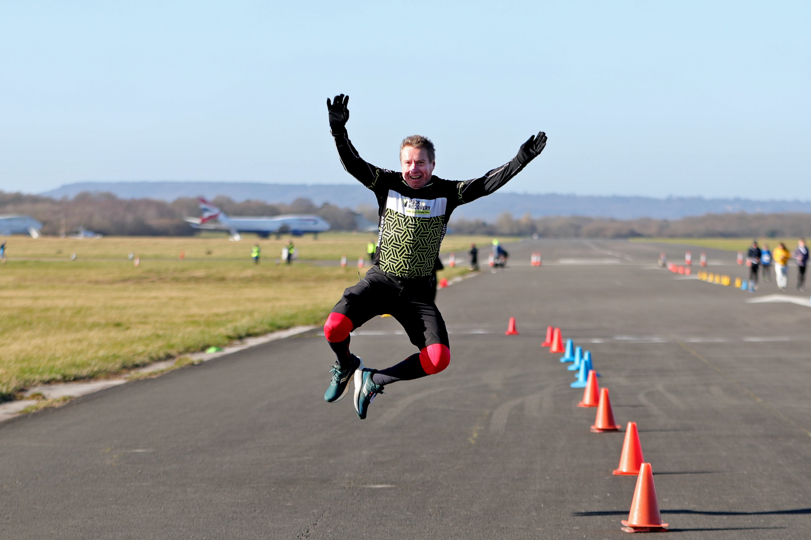 A person in athletic gear, including black and red tights and a green and black top, jumps joyfully with arms raised on an empty airstrip lined with orange and blue cones. Several airplanes and open grass fields are visible in the background.