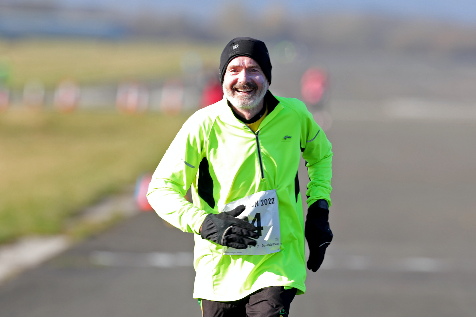 A man wearing a fluorescent yellow-green jacket, black gloves, and a black beanie runs on a paved road. He is smiling and has a race bib pinned to his jacket. The background is blurred, showing other runners and open fields in the distance.