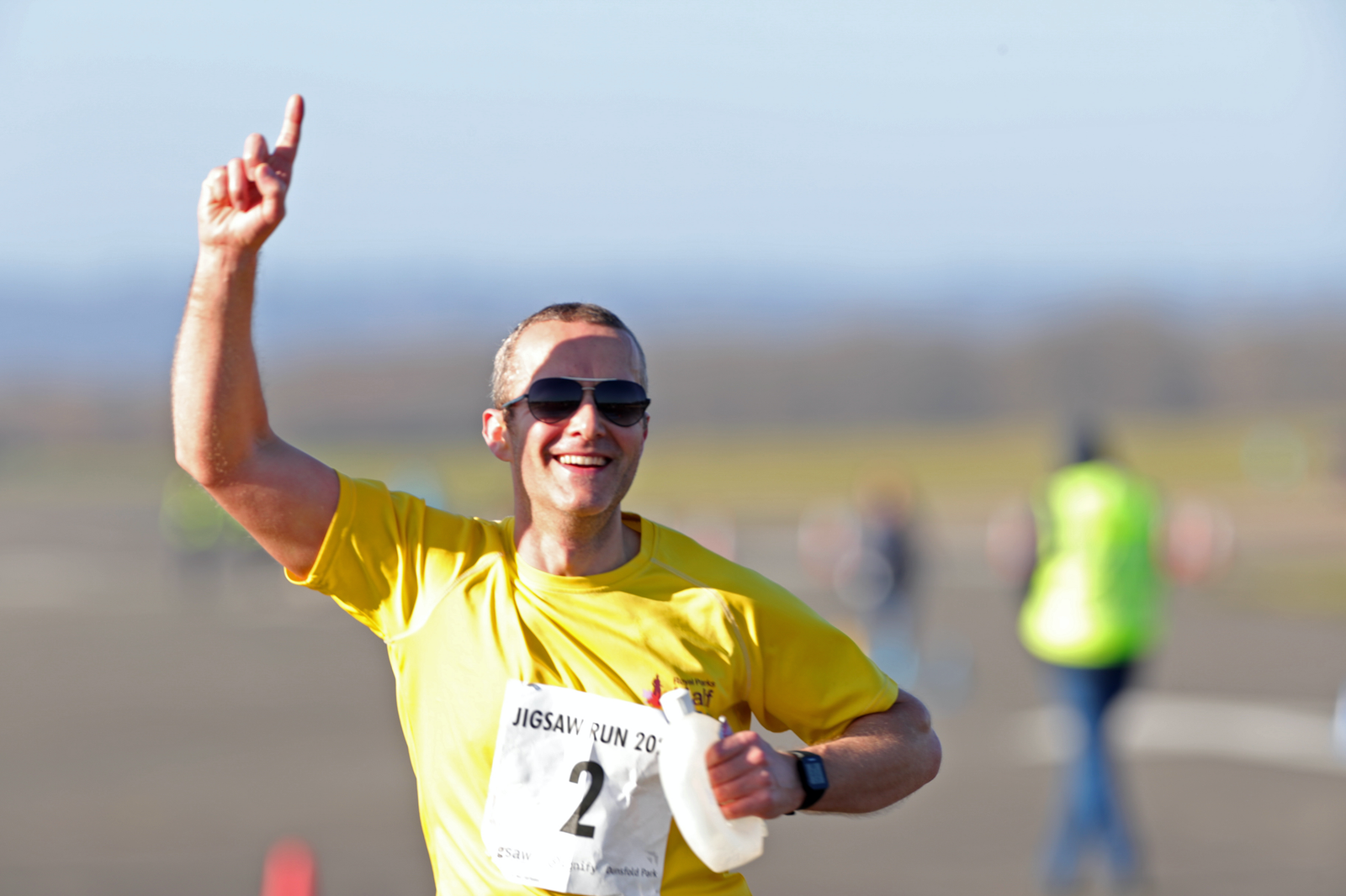 A person wearing a yellow shirt with the number 2 running in a race, smiling, and raising their left index finger. They are wearing sunglasses and there are blurred figures and an open field in the background.