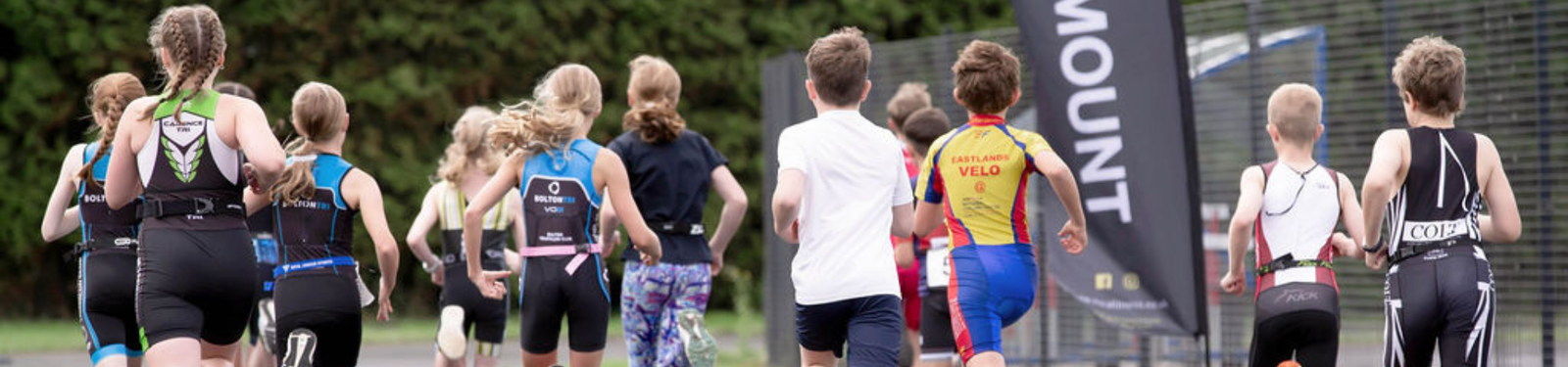 A group of children in athletic gear are running on a track during a race. They are wearing various colorful outfits, and a banner is visible in the background. Trees line the edge of the track.
