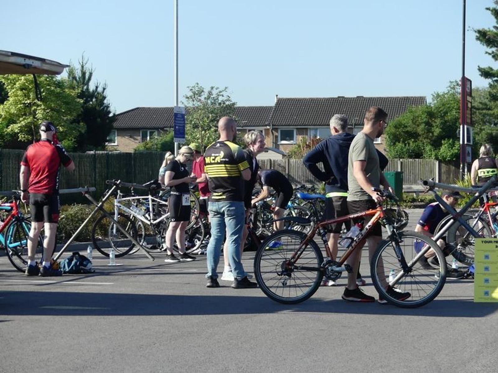 A group of cyclists gather in a parking area with their bikes. Some are adjusting equipment while others stand and chat. The setting is sunny, with trees and residential buildings in the background.