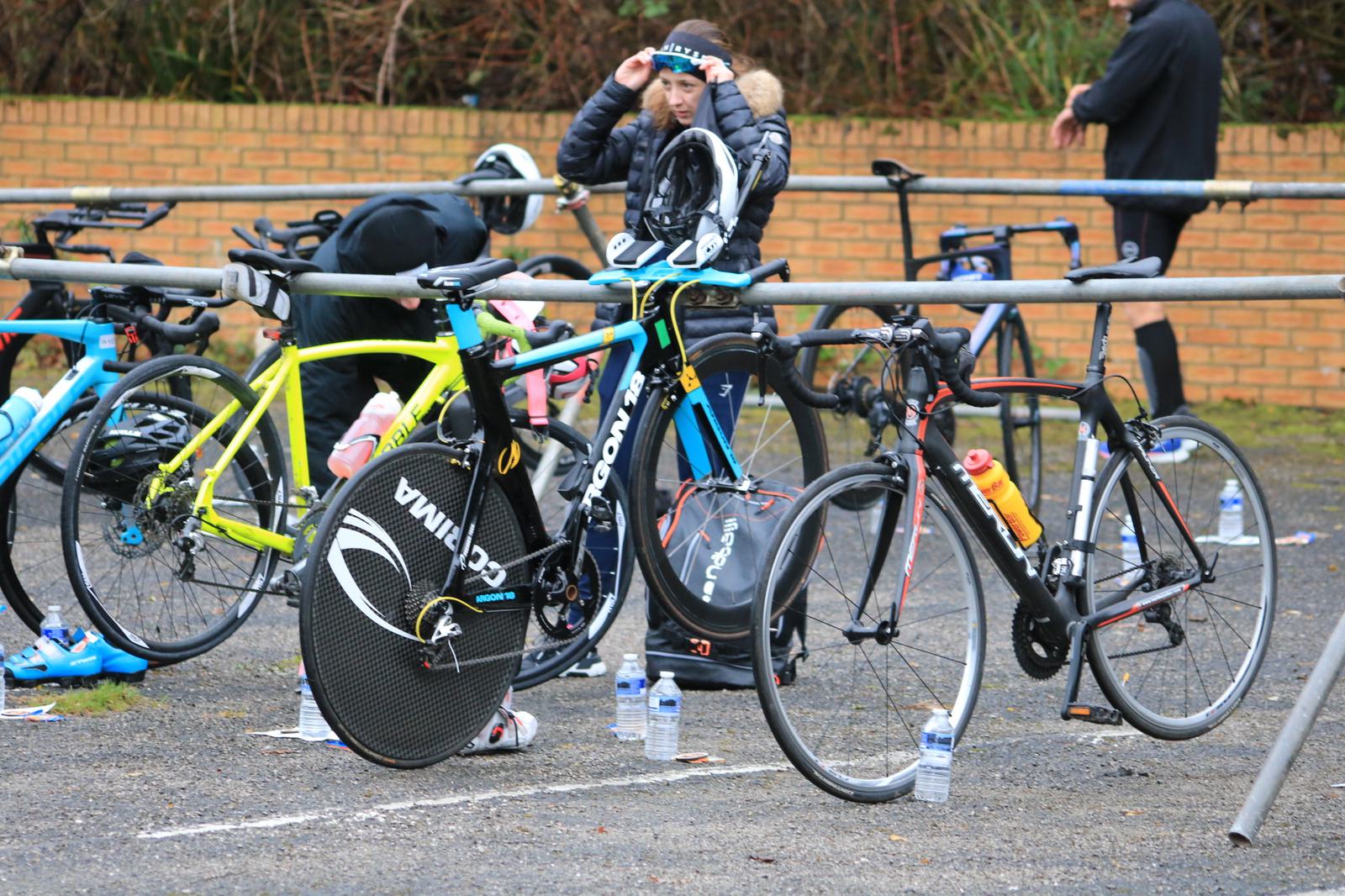 A row of bicycles is parked at a cycling event, each equipped with gear and water bottles. A cyclist adjusts their helmet in the background. The area is outdoors, with a brick wall and scattered water bottles on the ground.