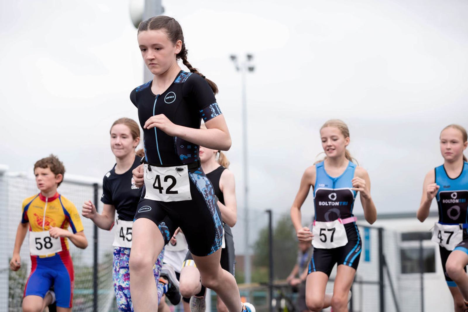 A group of young athletes running in a competitive race, wearing numbered bibs. The focus is on a girl with the number 42, surrounded by other participants. The background shows a cloudy sky and fencing.