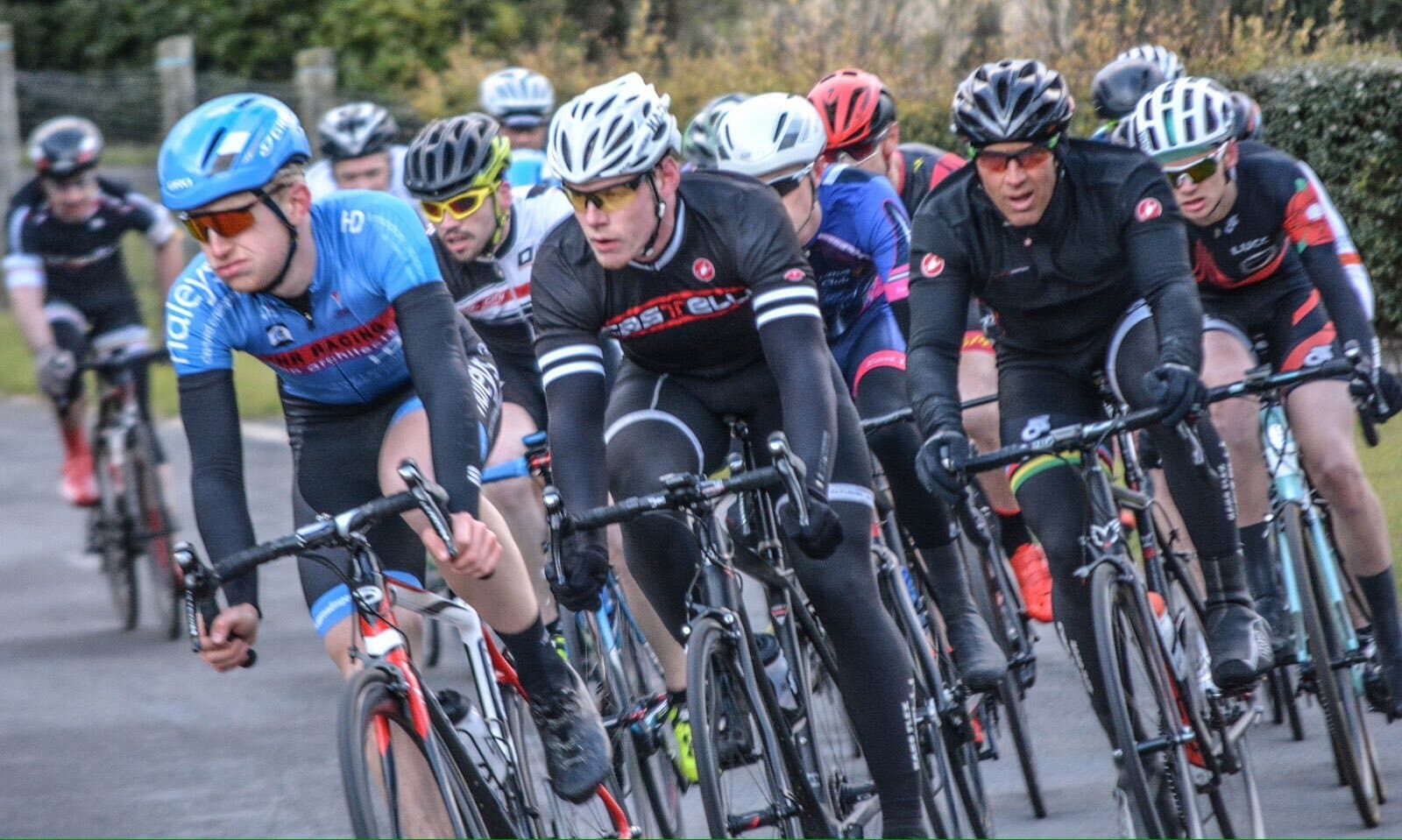 A group of cyclists wearing various uniforms and helmets race along a road. The cyclist in the front wears a blue jersey, while others behind don dark-colored outfits. Trees and a fence are blurred in the background, indicating their speed.