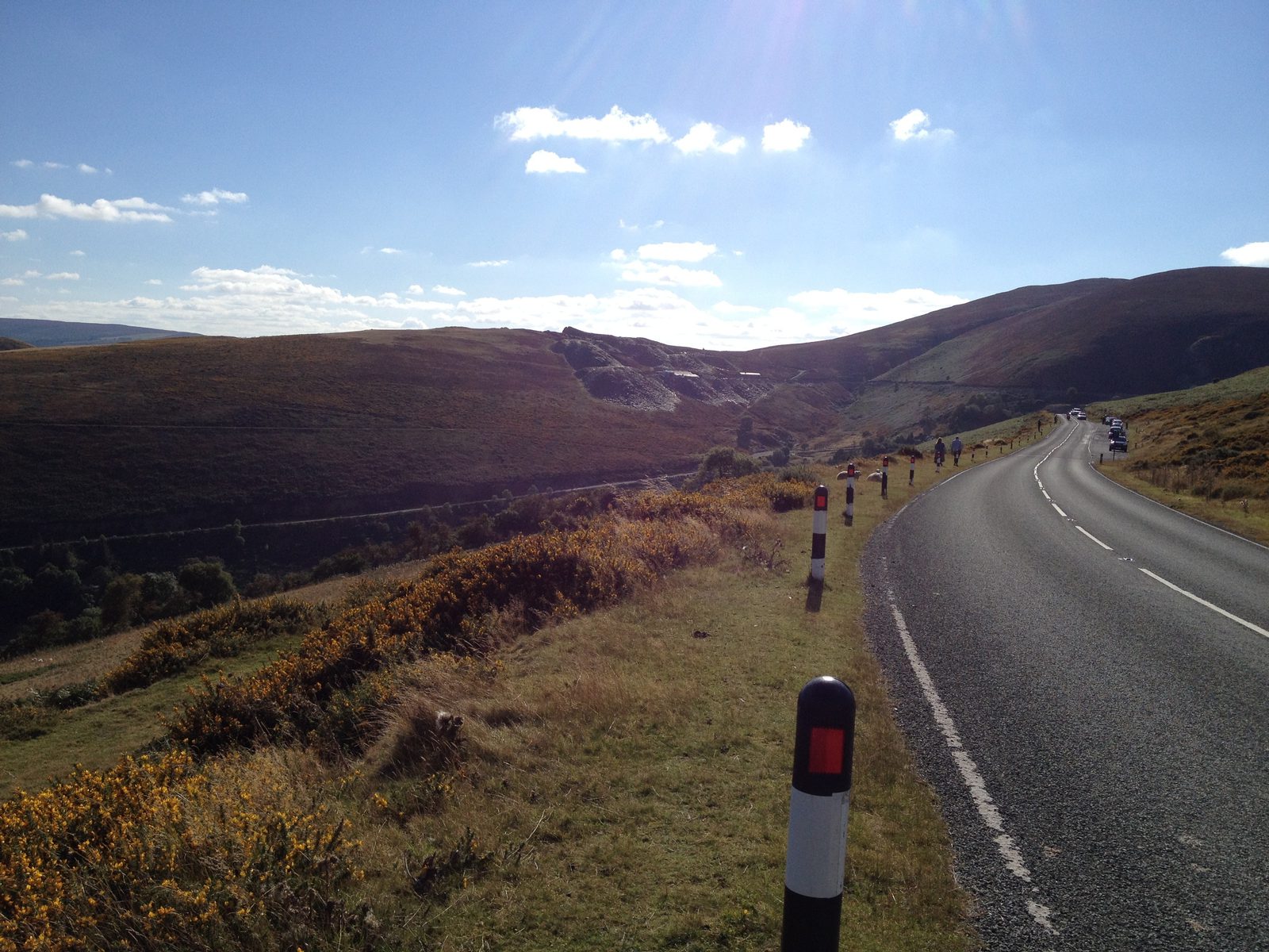 A winding road with black and white bollards leads through a hilly landscape covered in grassy and bushy vegetation. The sky is clear with scattered clouds, and the sun is shining brightly, creating a tranquil and scenic view.