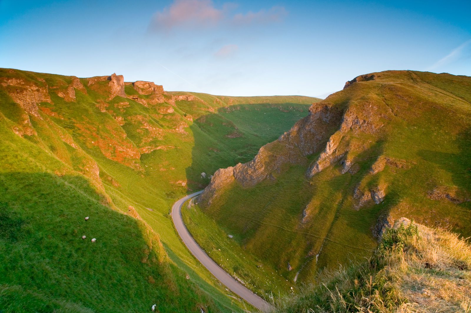 A winding road curving through a lush, green valley with steep, grassy hills on either side. The sky is clear with a warm, golden light during sunset, casting long shadows over the landscape and highlighting the rugged terrain.