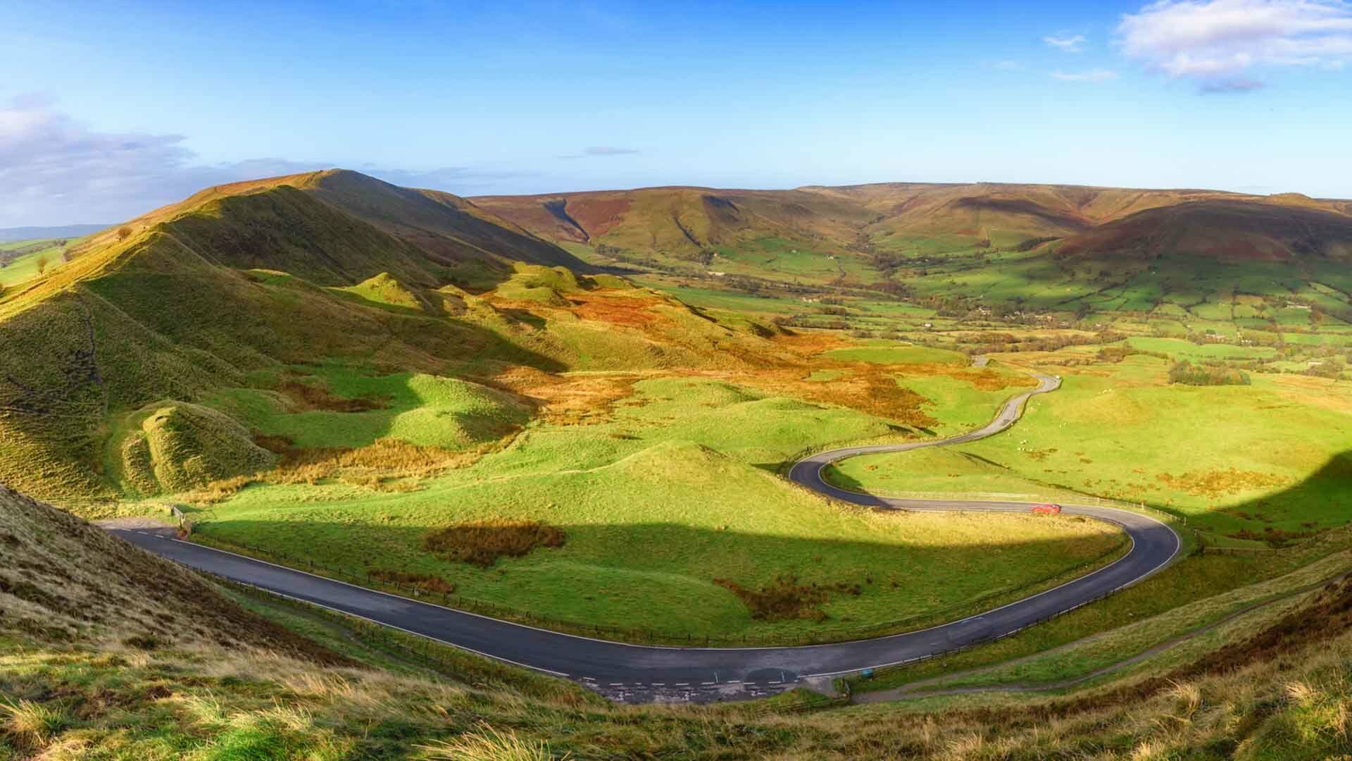 A winding road snakes through a lush green valley surrounded by rolling hills under a partly cloudy sky. Patches of golden brown vegetation dot the landscape, and shadows from the hills add depth to the scenic panorama.