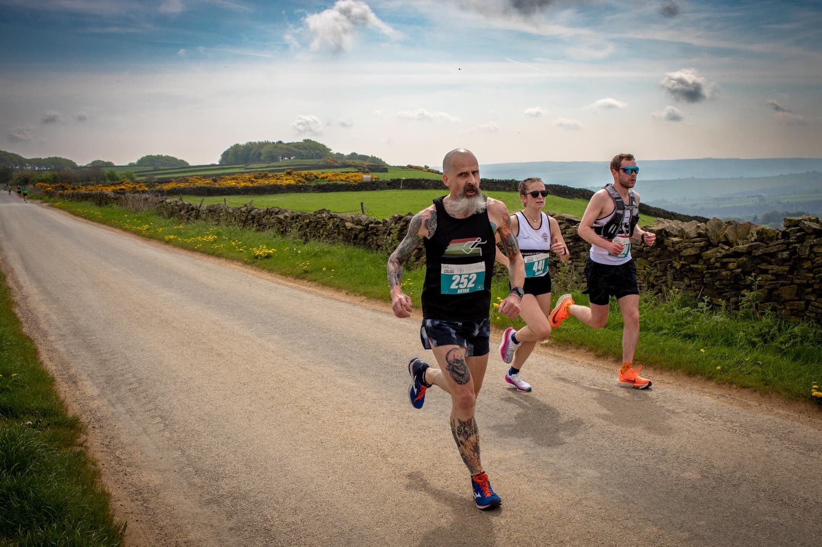 Three runners in athletic gear sprint down a rural road surrounded by fields and stone walls. The sky is partly cloudy, and the runners appear focused and determined.