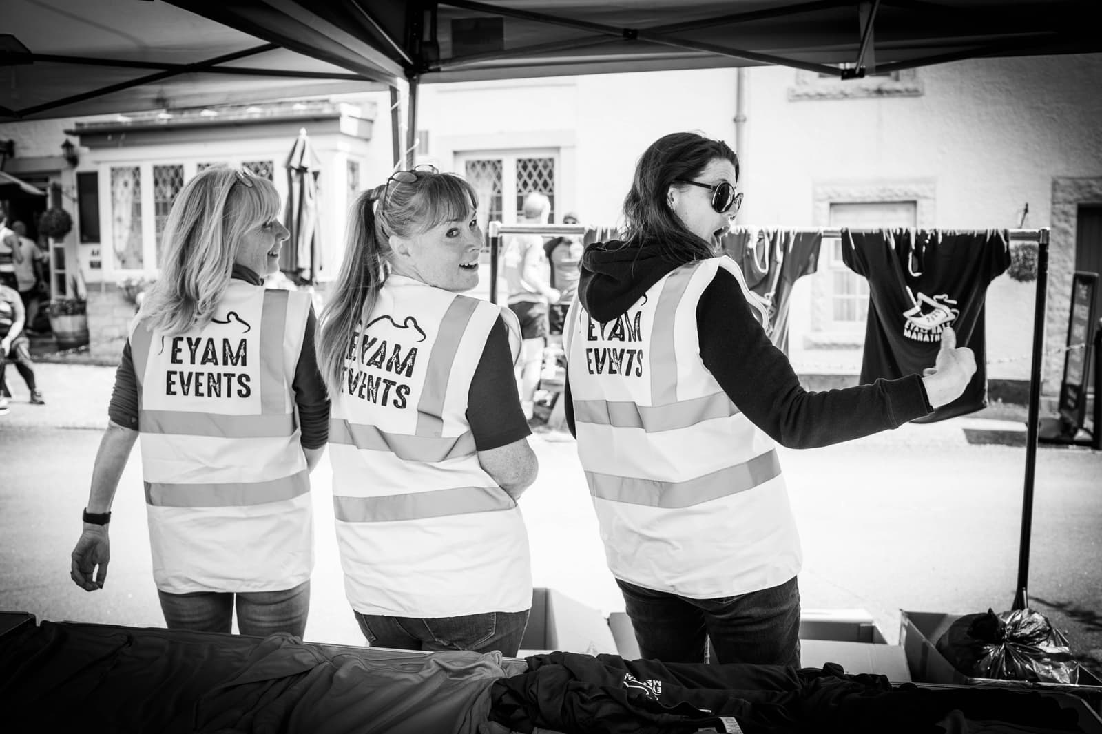 Three women wearing "Eyam Events" safety vests stand under a tent. One holds up a T-shirt while the others face the camera smiling. Clothing is displayed on tables and hanging nearby. Scene appears to be part of an outdoor event.