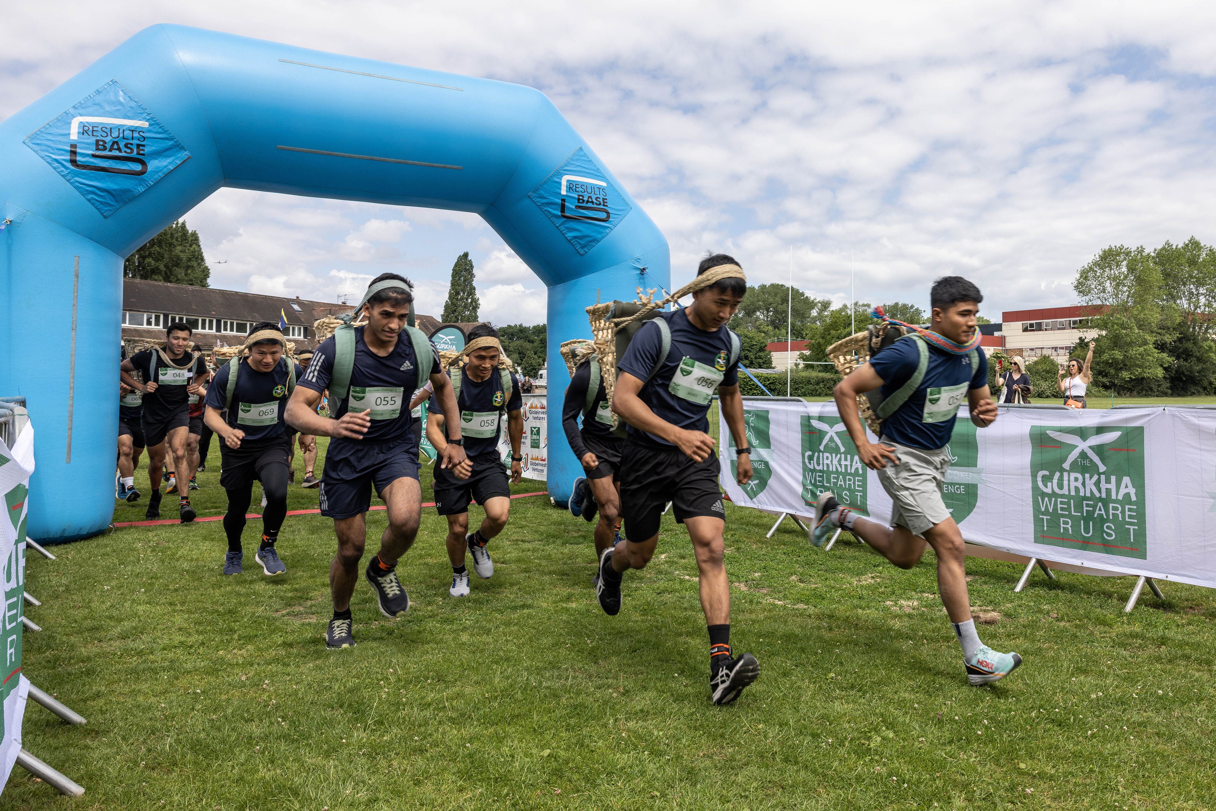 A group of runners starts a race, emerging from a blue inflatable arch with the words "Reservist Base" on it. They wear numbered bibs and athletic gear. The event appears to be organized by or in support of the Gurkha Welfare Trust, as indicated by signs. Trees and buildings are in the background.