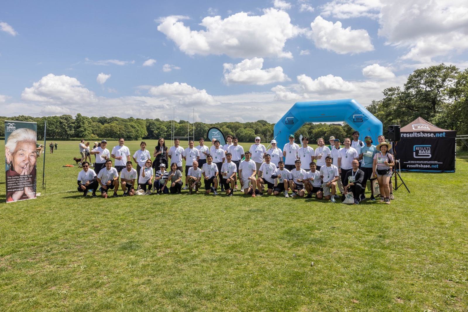 A large group of people, all wearing white T-shirts, are posing for a photo on a grassy field under a partly cloudy sky. Behind them are a blue inflatable arch, some trees, and promotional banners. They appear to be part of an outdoor event or race.