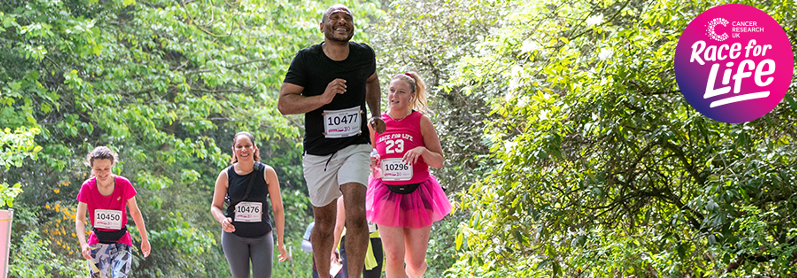 Four participants in athletic attire are running on a tree-lined path during a Race for Life event. The foreground runner, wearing a numbered bib and black shirt, strides ahead, while a woman in a pink tutu follows. The event logo is in the top-right corner.