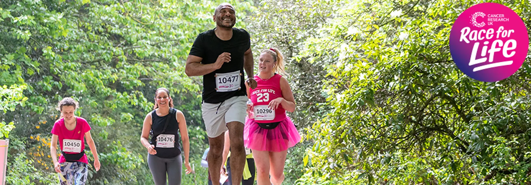 A group of people run in a charity event through a lush, green pathway. They wear race numbers and themed pink clothing with a "Race for Life" logo in the corner.