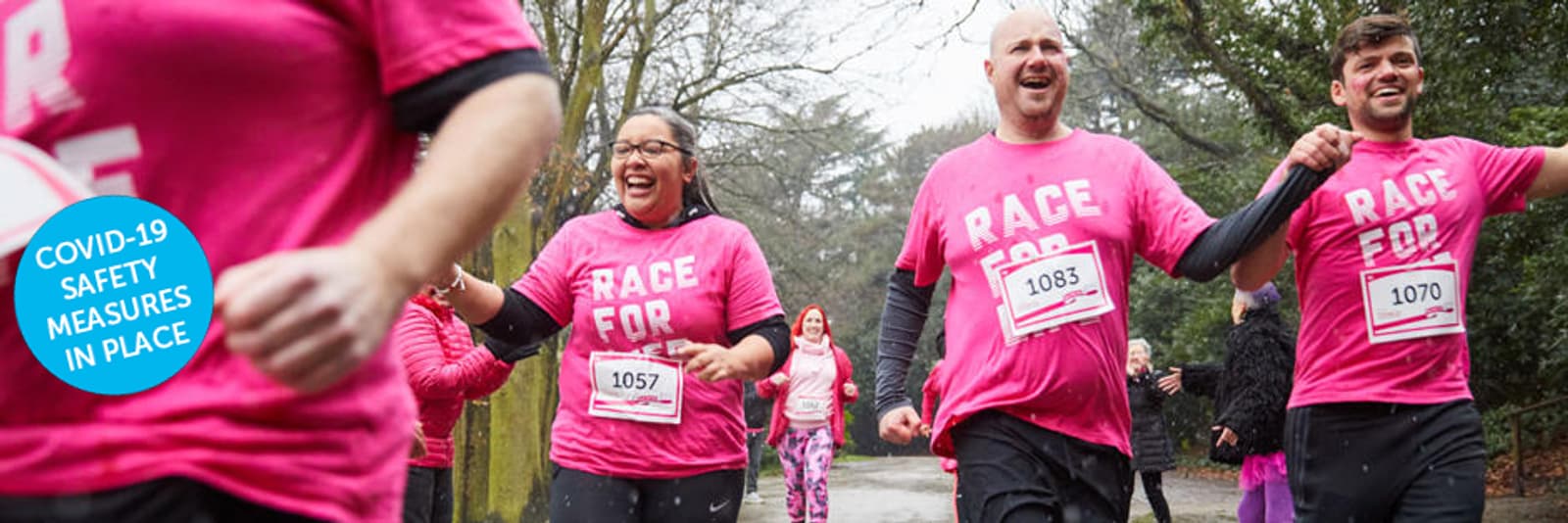 A group of happy runners wearing matching pink shirts with "RACE FOR" printed on them run outdoors on a tree-lined path. They are all smiling and appear to be participating in an organized race. A blue circle on the left reads "COVID-19 SAFETY MEASURES IN PLACE.