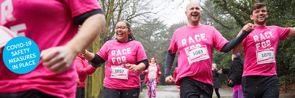 A group of people wearing bright pink shirts that read "Race For" participate in an outdoor run. They are smiling and holding hands as they move forward. Bib numbers 1057, 1083, and 1070 are visible. A blue sign reads "COVID-19 Safety Measures in Place.