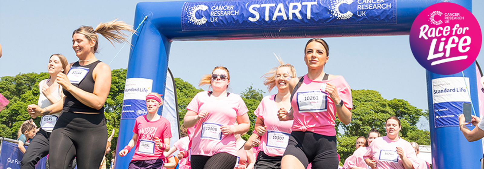 A group of women in pink shirts and athletic wear participate in a Race for Life event, running and smiling as they pass under a blue "START" archway. Trees are visible in the background, and the Race for Life logo is in the top right corner.