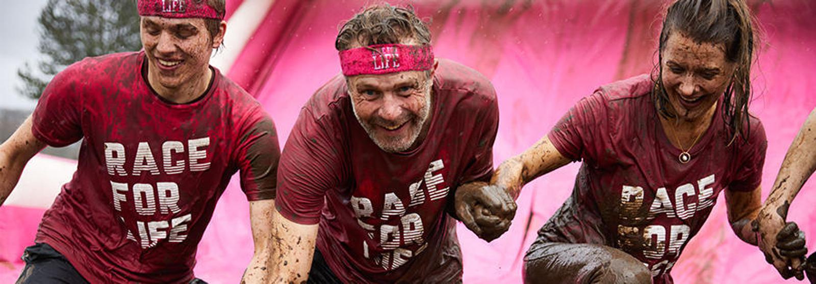 Three people in muddy red "Race for Life" shirts and headbands smile as they navigate a slippery, mud-covered obstacle. They appear to be enjoying the challenge and supporting each other through the race. The background is dominated by a large pink inflatable slide.