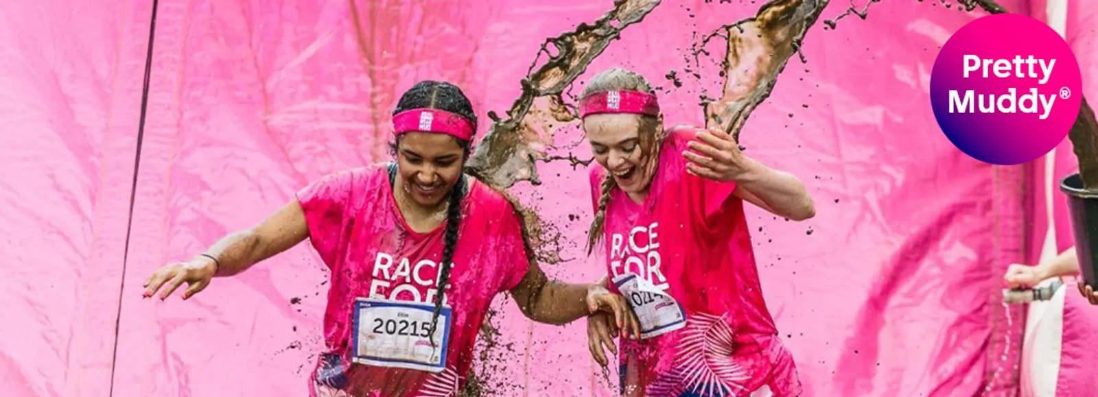 Two people in pink shirts and headbands run through a mud obstacle, laughing as mud splashes on them. The event is part of "Pretty Muddy." They wear race numbers and the backdrop is a pink inflatable structure.