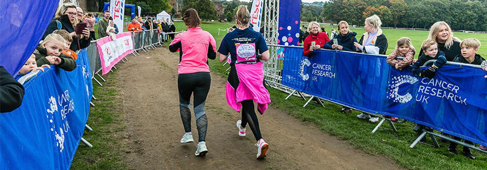 Two runners, one in pink and the other in black with a pink jacket tied around the waist, approach the finish line at a charity event. Spectators line the route, cheering. Banners and flags with the "Cancer Research UK" logo are visible.