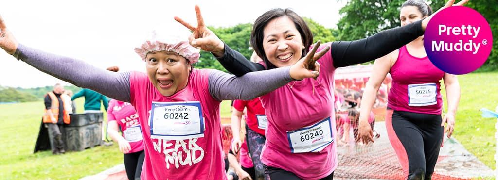Two women, wearing pink shirts and race bibs, are smiling and posing with outstretched arms during a "Pretty Muddy" obstacle race. Other participants are visible in the background on a grassy field. A "Pretty Muddy" logo is in the top right corner.