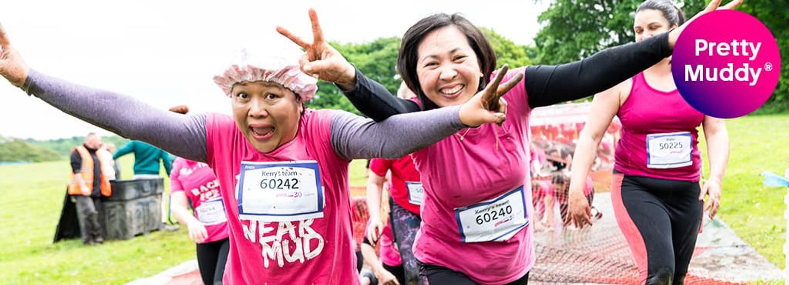 Two women, wearing pink shirts and race bibs, are smiling and posing with outstretched arms during a "Pretty Muddy" obstacle race. Other participants are visible in the background on a grassy field. A "Pretty Muddy" logo is in the top right corner.