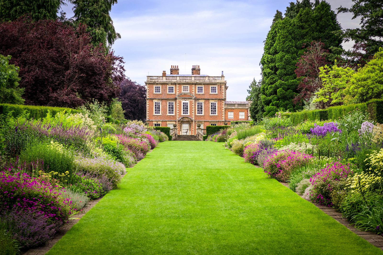A stately brick mansion with symmetrical rows of windows and chimneys stands at the end of a lush, manicured green lawn. The lawn is flanked by vibrant, colorful flower beds and neatly trimmed hedges, with tall trees in the background under a partly cloudy sky.