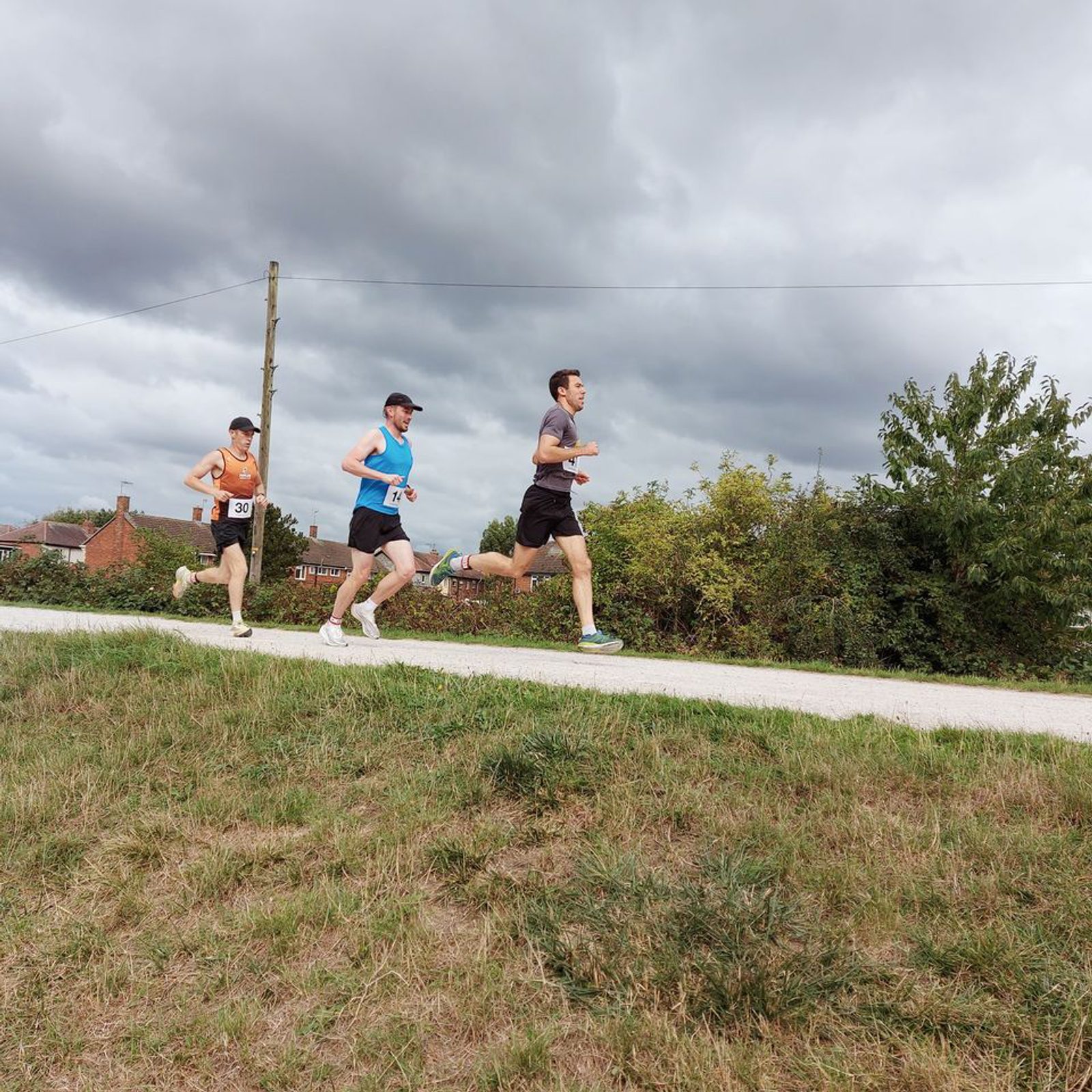 Three individuals jogging on a gravel path surrounded by greenery under a cloudy sky. The first person wears an orange top, the second wears a blue top and white cap, and the third wears a gray top. They are mid-stride, with houses visible in the background.