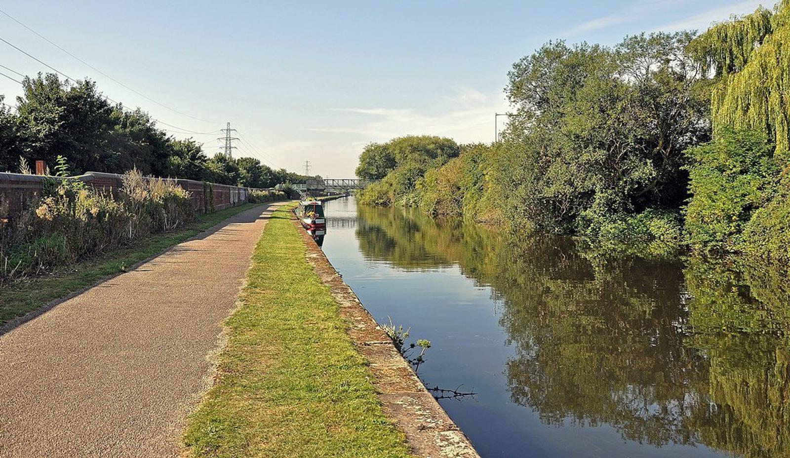 A calm canal flanked by lush greenery on one side and a paved pathway on the other. A lone person in a small boat floats on the water. The sky is clear blue, and a bridge is visible in the distance.