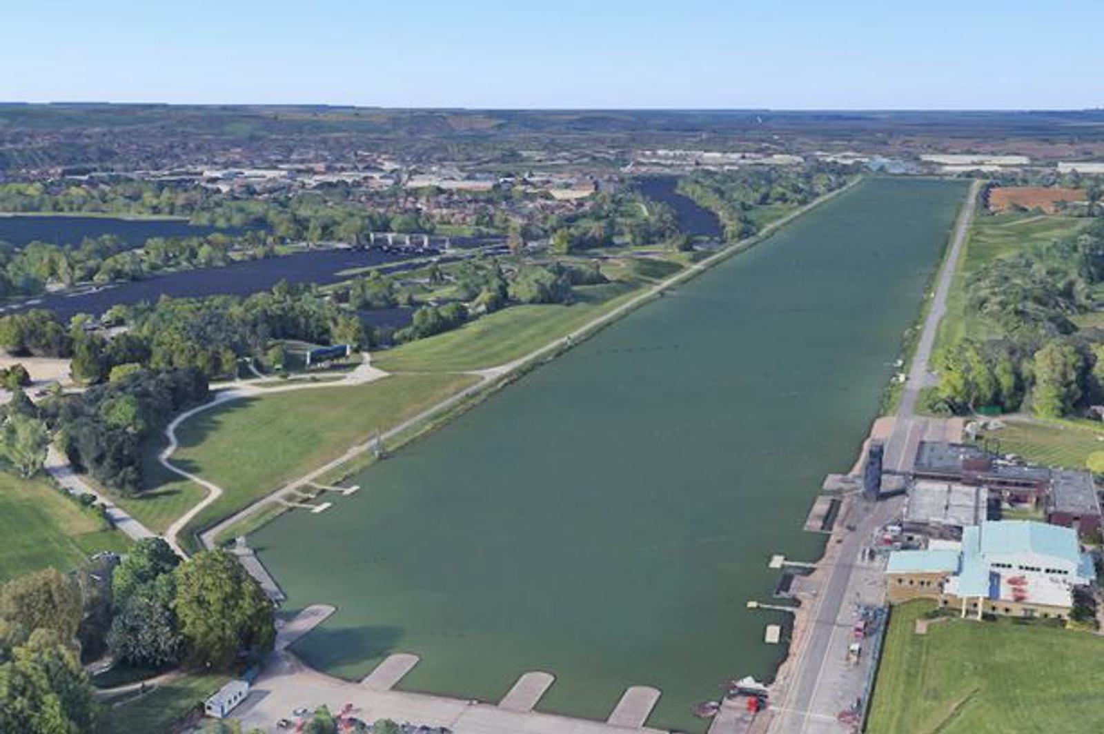 Aerial view of a long, narrow rowing lake surrounded by green fields and trees. Adjacent to the lake are pathways and a few buildings. In the distance, a town and hilly landscape are visible under a clear sky.