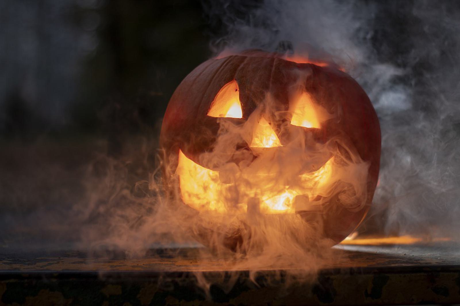 A carved pumpkin lantern, commonly known as a jack-o'-lantern, glows from within. The pumpkin has a spooky face with triangular eyes and a wide, toothy grin. Thick mist or smoke surrounds it, enhancing the eerie atmosphere. The background is dark and blurred.