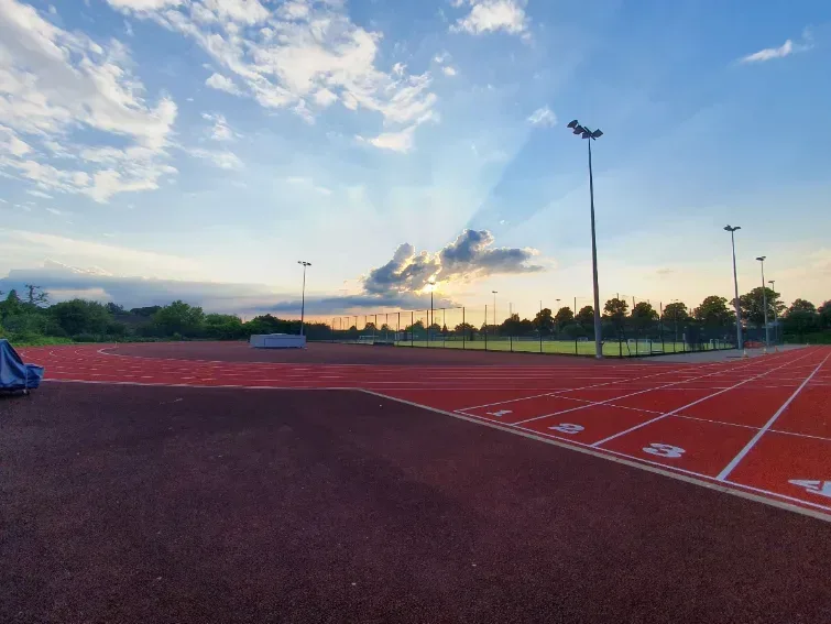 A running track under a sky with scattered clouds at sunset. The track is empty, with lane numbers visible in the foreground. There are floodlights around the track, and trees and vegetation can be seen in the background.