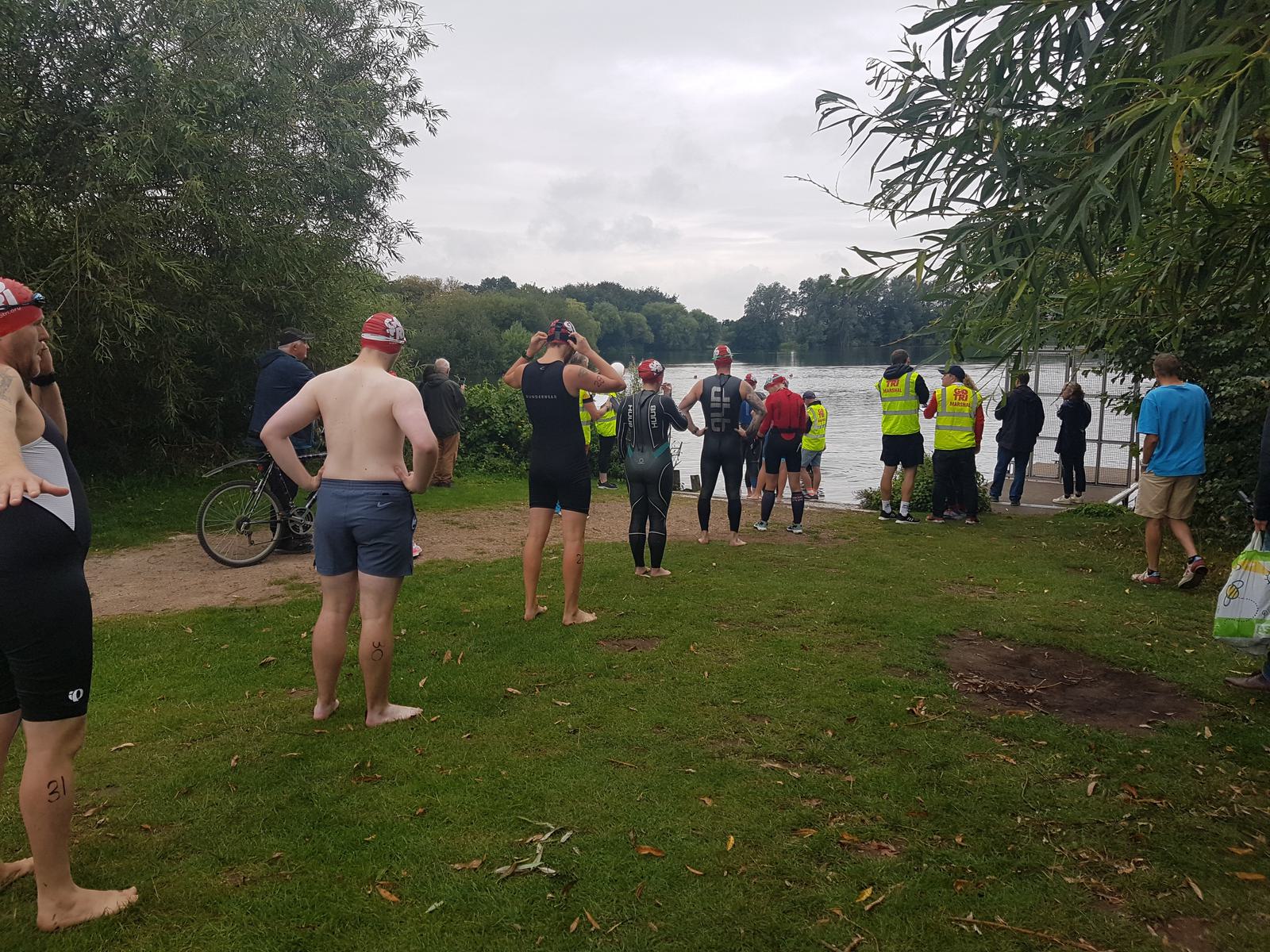 Participants in wetsuits and red swim caps gather at the edge of a lake, preparing for a swim event. Spectators and officials in yellow vests stand nearby. The scene is set in a grassy area with trees, a cloudy sky overhead, and a bicycle in the background.