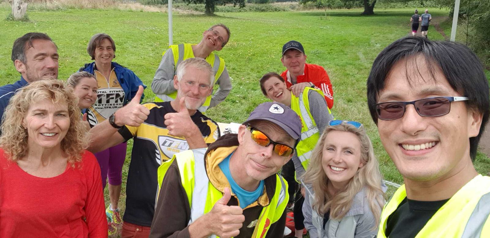 A group of people, most in high-visibility vests, smile and pose for a photo outside on a grassy area with trees in the background. One man in the center gives a thumbs-up, while another at the front wears sunglasses and a cap.