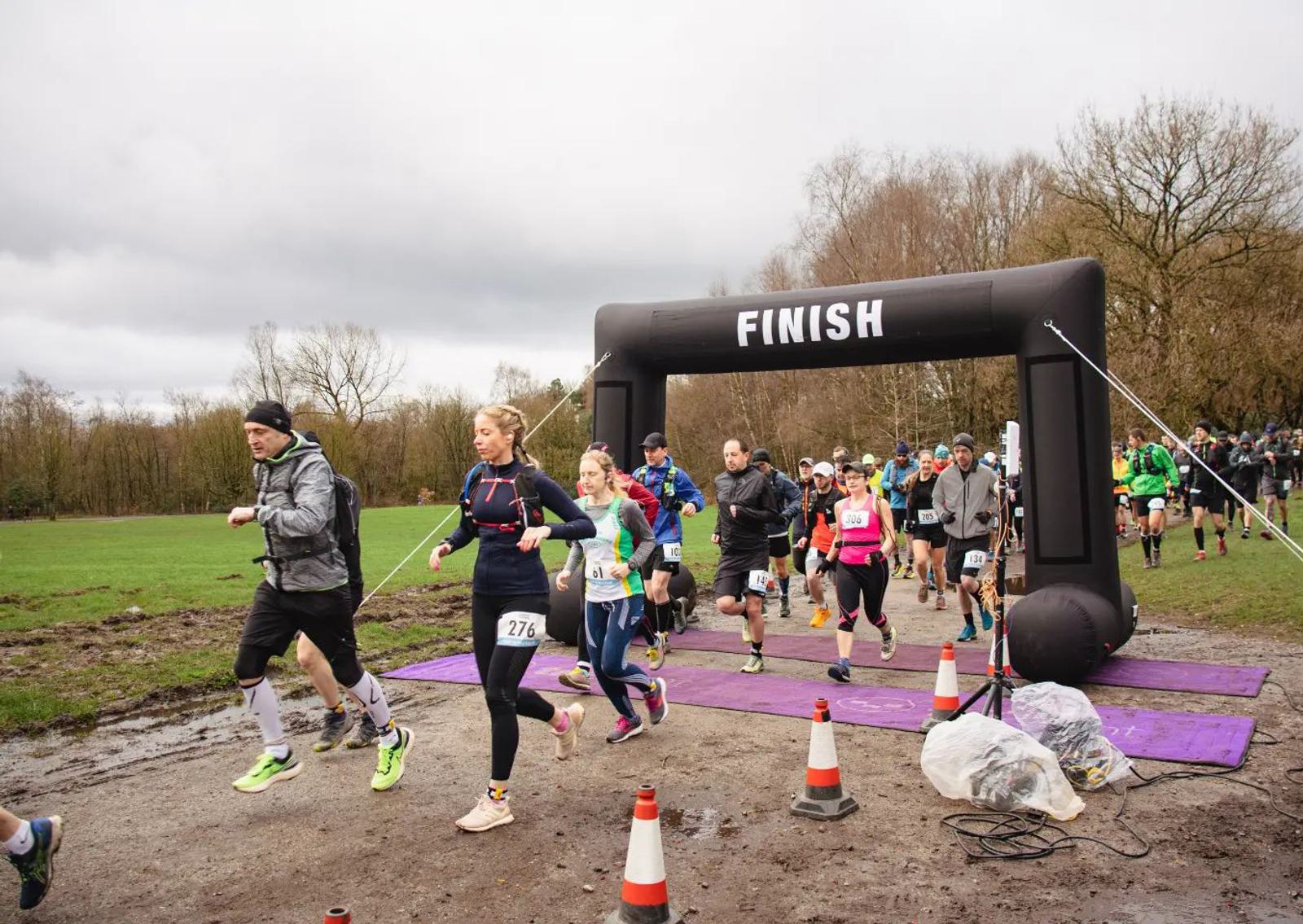 A group of runners is crossing the start line of a race on an overcast day. The runners, dressed in varied athletic gear, are moving through a muddy path with trees in the background. Orange cones and a large "FINISH" arch are visible, surrounded by spectators.