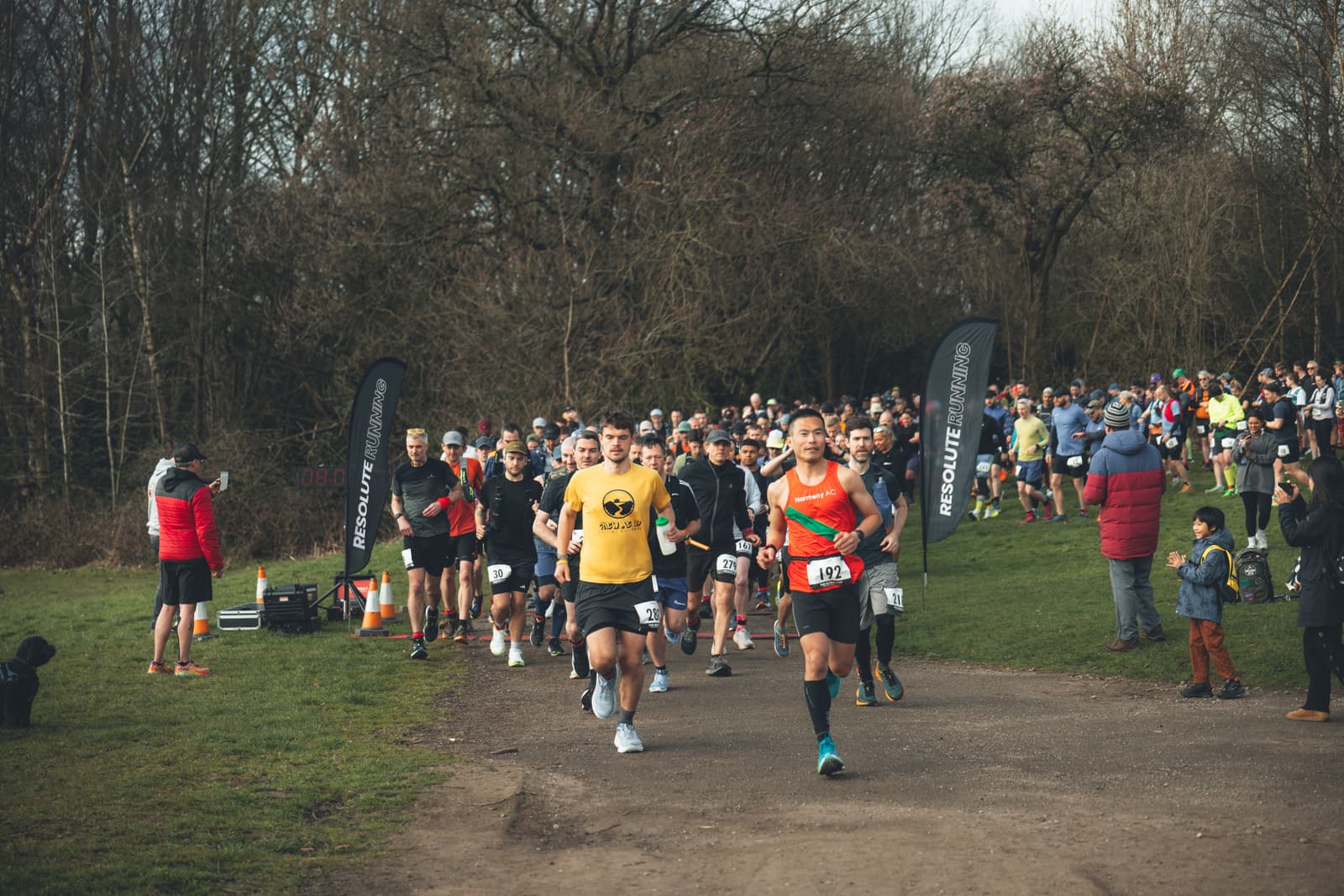Runners participating in a race on a path through a park lined with trees. They are surrounded by spectators and event banners. One runner in a yellow shirt leads the group, while another in an orange top follows closely.