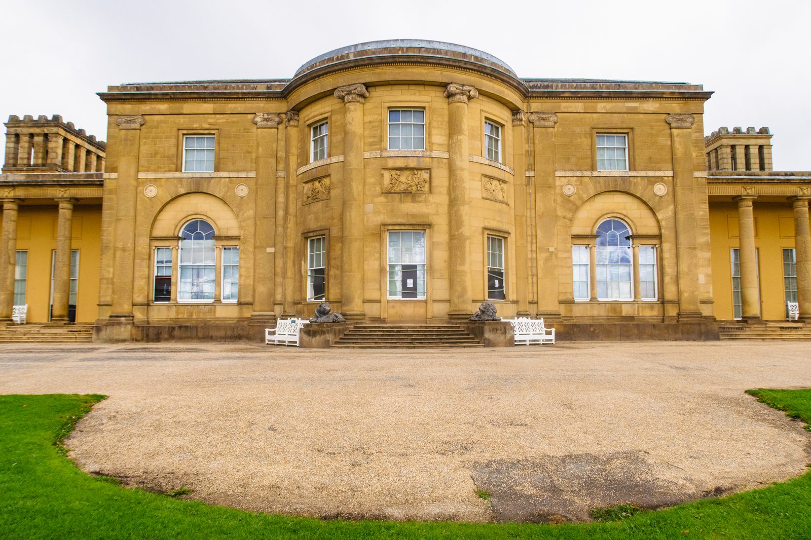 Front view of a grand, historic building with symmetrical architecture and stone facade. Features three arched windows and an ornate central section with columns. Two white benches and two lion statues are placed near the entrance steps. The ground is gravelly.