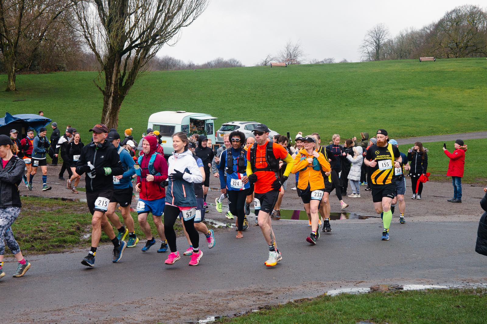 A group of runners in colorful athletic wear participate in a race on a cloudy day in a park. Some trees and a green grassy hill are visible in the background, along with a white camper van parked nearby. Spectators and participants gather along the route.