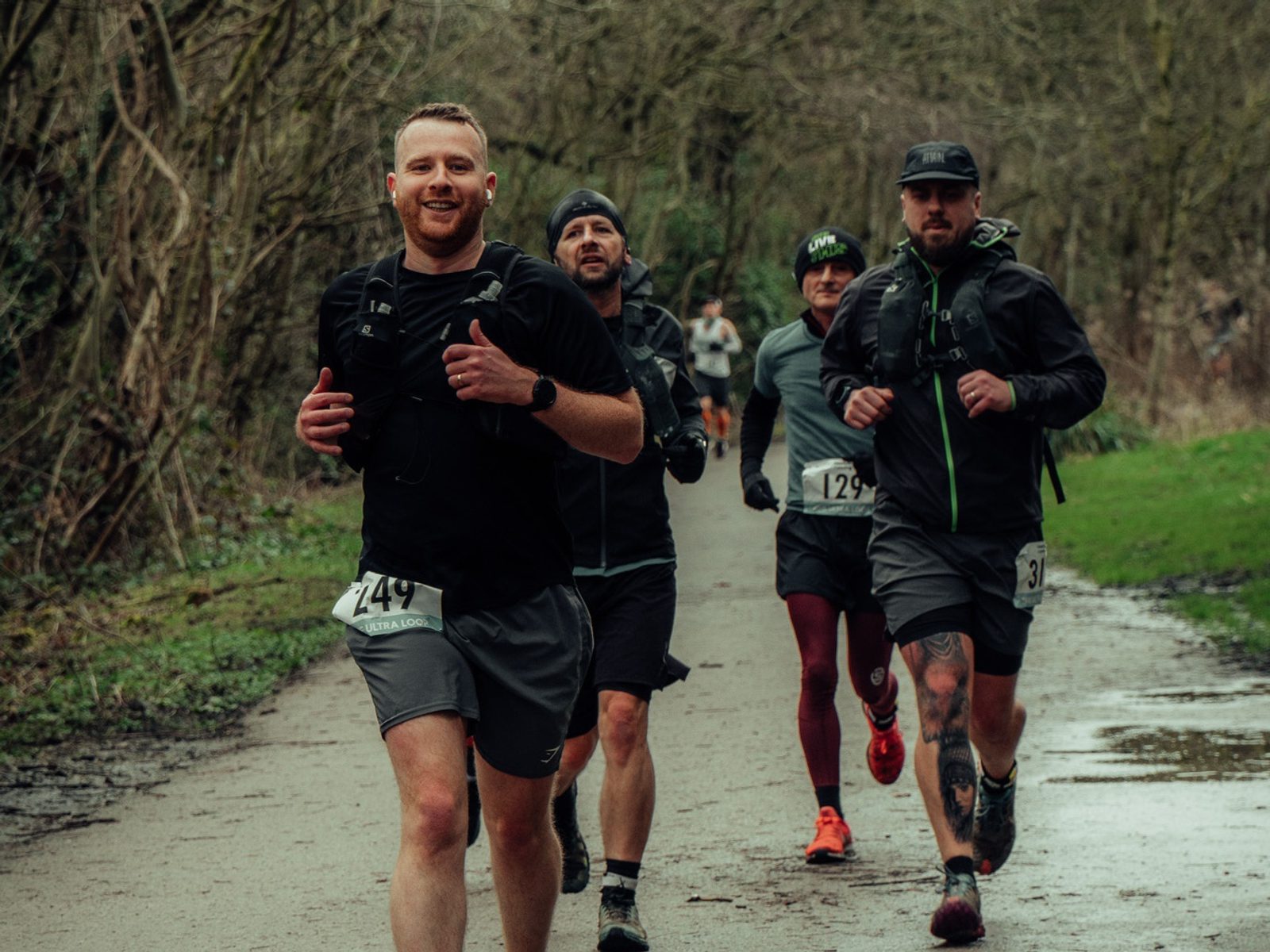 Four runners are participating in an outdoor race along a trail surrounded by trees. The runner in front wears a black shirt and smiles at the camera, while the others follow closely behind, all dressed in activewear and carrying gear. The ground is slightly wet.