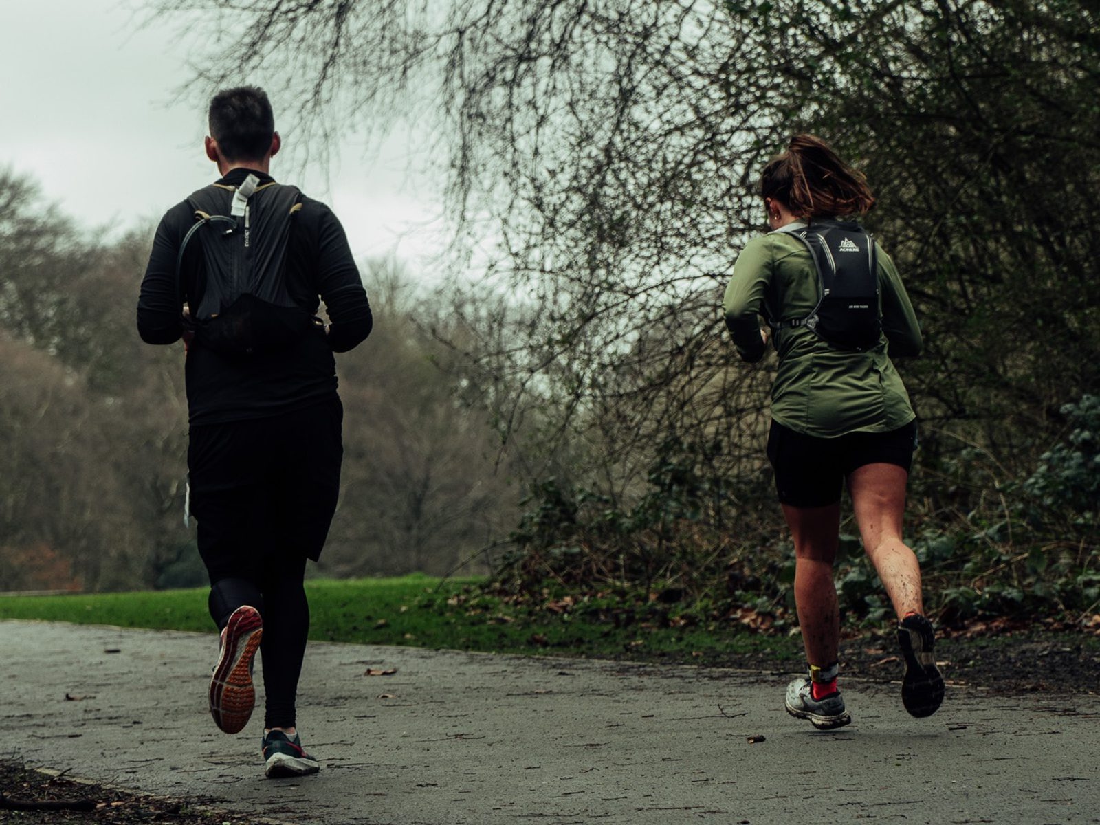 Two people jog on a path through a park, both wearing dark athletic clothing and backpacks. The path is wet and surrounded by leafless trees and shrubs, indicating a cool, overcast day. One runner wears black shorts, the other is in long black leggings.