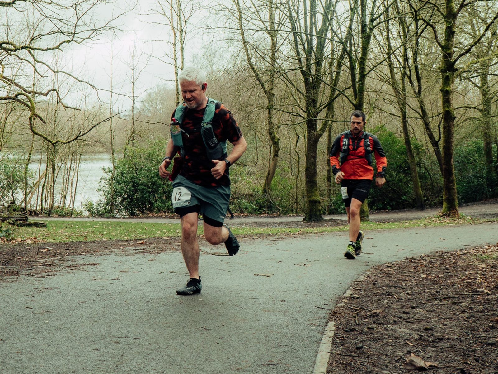 Two men are running along a path in a forested park. The man in front is wearing a dark shirt and shorts, while the man behind him is wearing a red jacket and black shorts. Both are carrying small backpacks and have racing bibs pinned to their clothing. Trees surround the path.