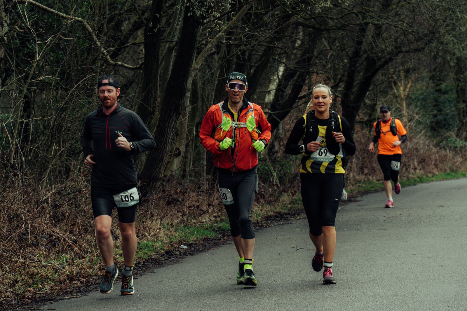 Four runners compete in a marathon on a forested road. The group includes three men and one woman. Two runners are wearing numbered race bibs (106 and 69), one wears a red jacket, and the woman wears a black and yellow top. Trees line the path they are running on.