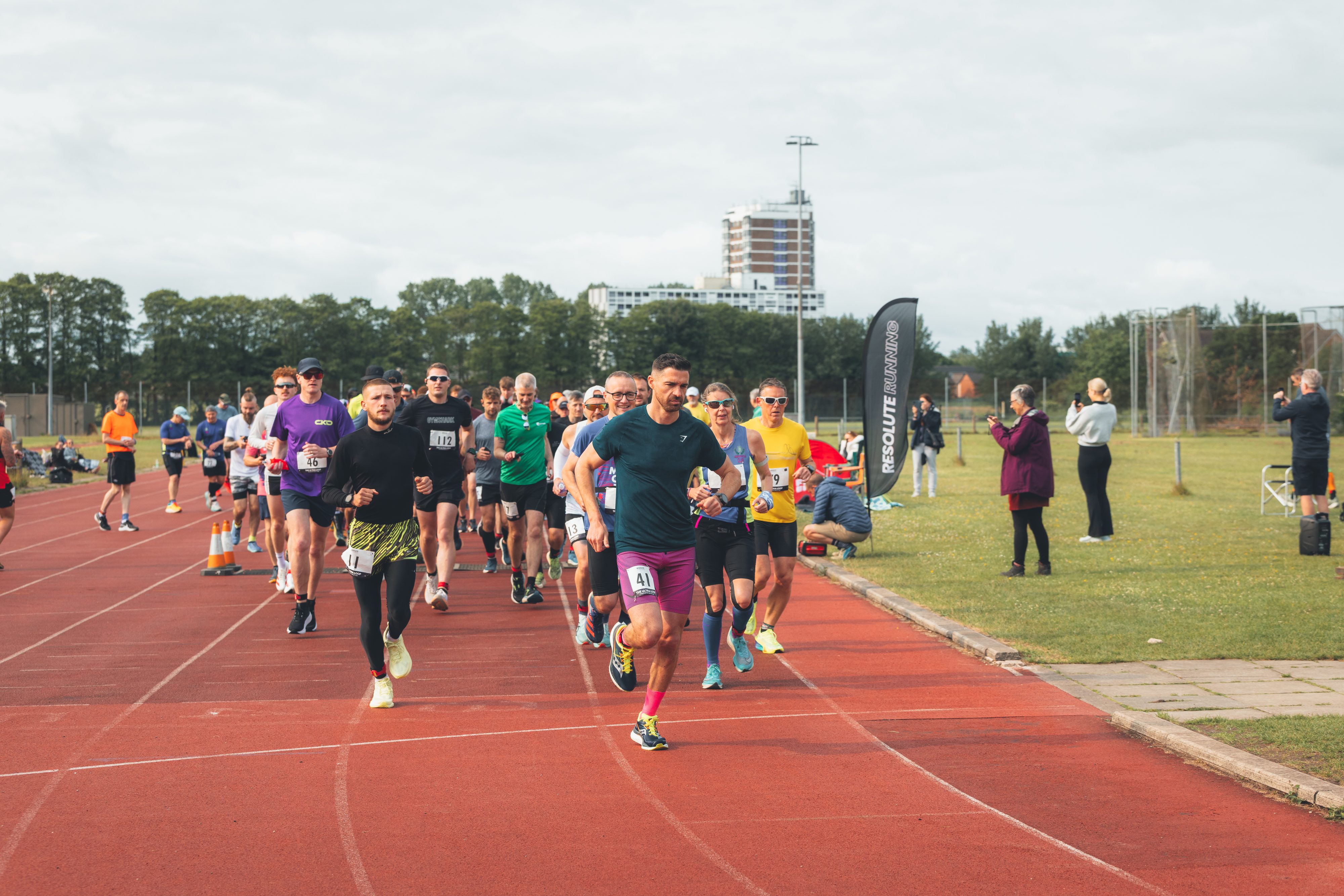 Runners in athletic wear participate in a race on a red track, with spectators and event staff watching from the sidelines. Trees and a tall building are visible in the background under a cloudy sky.