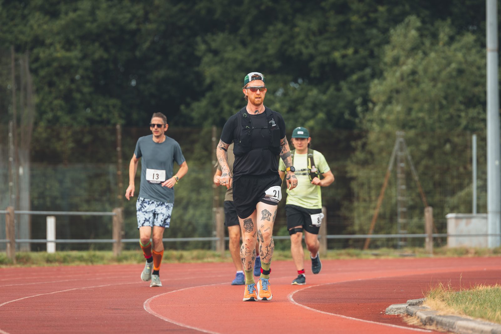 Four men participate in a race on an outdoor track, wearing race bibs and athletic gear. The man in front has tattoos and wears a cap and sunglasses, while the others follow behind him. Trees are visible in the background.